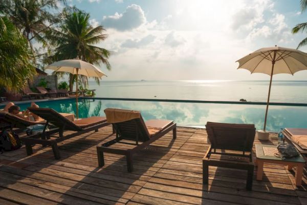 A serene poolside setting with loungers, umbrellas, and a view of the ocean, surrounded by palm trees under a bright sky.
