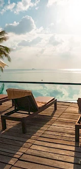Lounge chairs and umbrellas on a wooden deck by an infinity pool, overlooking a serene ocean under a partly cloudy sky.