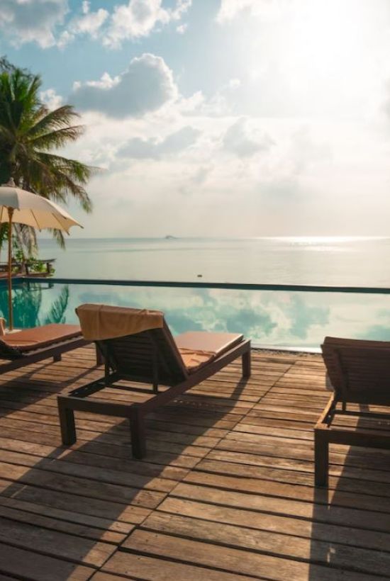 A serene poolside setting with lounge chairs, umbrellas, palm trees, and a stunning view of the ocean under a bright sky.