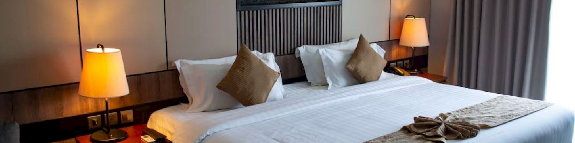 A modern hotel room features a large bed, bedside tables with lamps, and wall art. Curtains cover the window for privacy.
