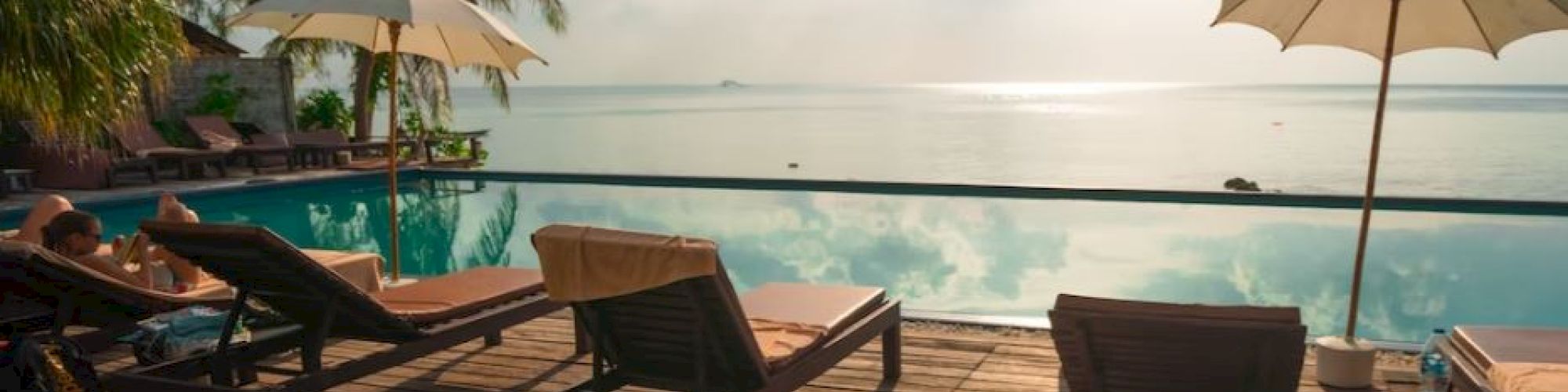 A serene poolside setting with lounge chairs, umbrellas, and palm trees overlooking a calm body of water under a partly cloudy sky.