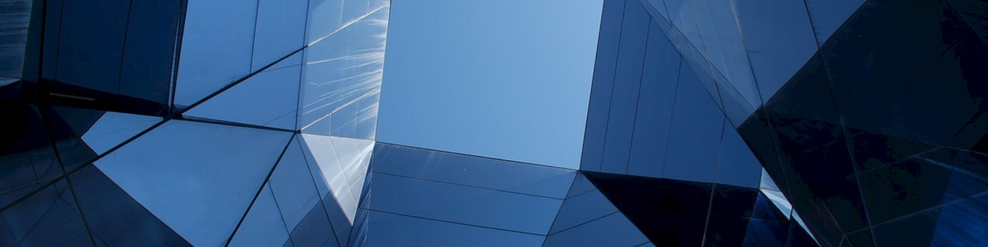 Geometric, mirrored architectural structure with blue glass panels forming a skylight, reflecting light and sky.