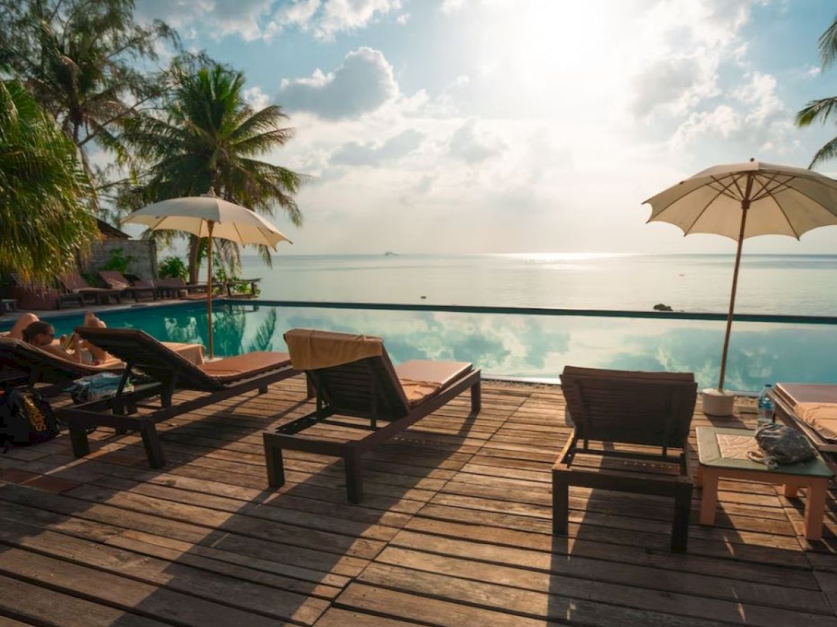 A serene poolside area with lounge chairs, umbrellas, palm trees, and a view of the ocean under a partly cloudy sky.