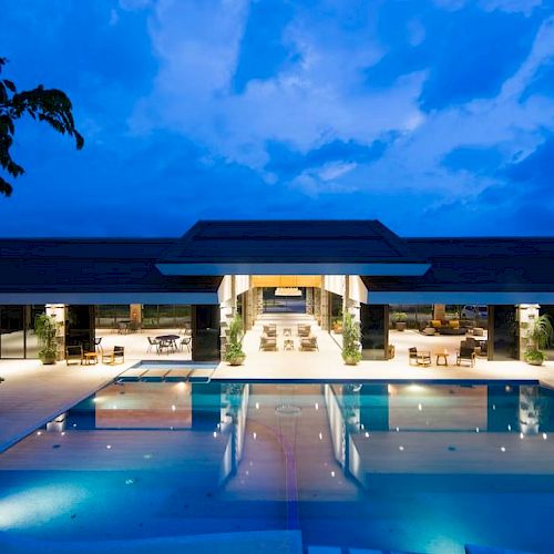 A luxurious outdoor pool area at night, featuring modern architecture, lit interiors, and a tranquil ambiance under a cloudy sky.