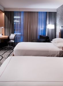 The image shows a modern hotel room with two beds, a desk, chair, lamps, and a wall-mounted TV. The room has stylish decor and neutral tones.