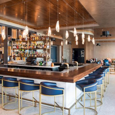 A modern bar with a stocked shelf, stylish pendant lights, and blue cushioned bar stools arranged around a wooden counter.