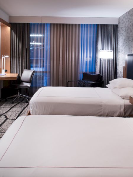 A modern hotel room with two beds, a TV, desk, and lamp. The design is sleek with patterned wallpaper and a large window with curtains.