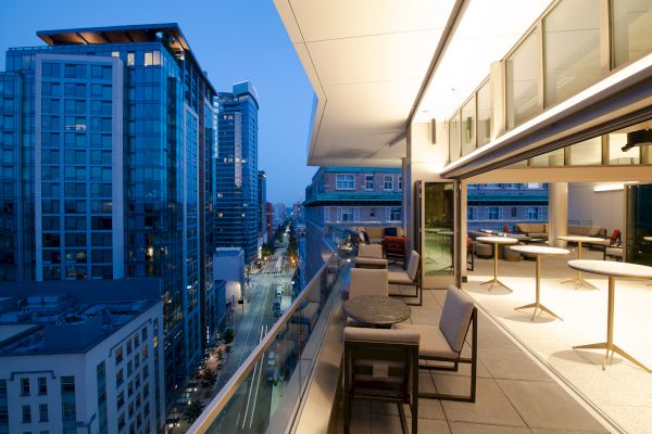 A modern rooftop terrace with tables and chairs overlooks a city street, surrounded by tall buildings in the evening light.