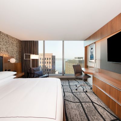 A modern hotel room with a bed, work desk, TV, and city view through large windows is shown.