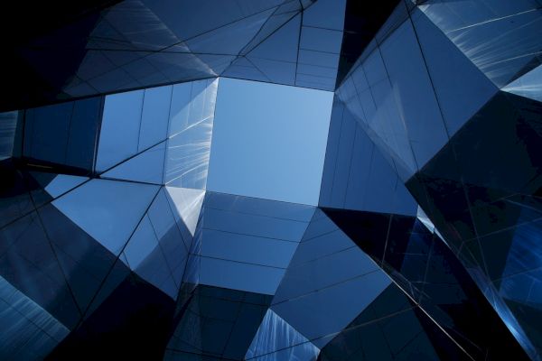 The image shows an upward view of modern glass architecture with geometric shapes and reflections under a blue sky.
