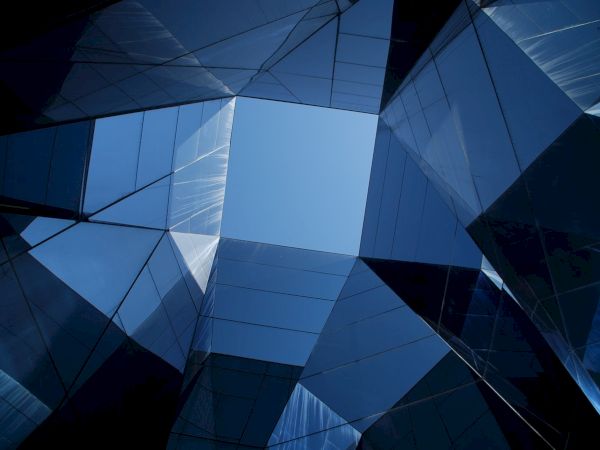 The image shows an upward view of modern glass architecture with geometric shapes and reflections under a blue sky.