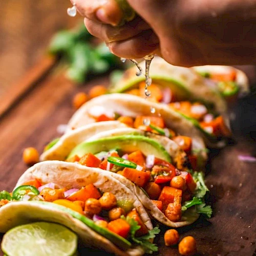 Vegetable tacos with chickpeas, avocado, and cilantro, topped with lime juice squeezed by a hand, arranged on a wooden board.
