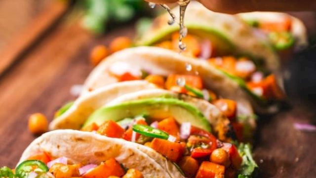 Vegetable tacos with chickpeas, avocado, and cilantro, topped with lime juice squeezed by a hand, arranged on a wooden board.