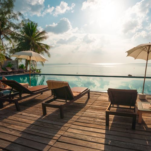 Sunlit poolside scene with lounge chairs, palm trees, and umbrellas overlooking a tranquil ocean view under a partly cloudy sky.