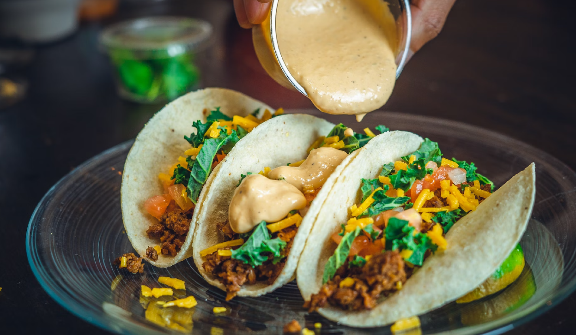 Three tacos with toppings like lettuce, cheese, and sauce being poured on them, set on a glass plate.