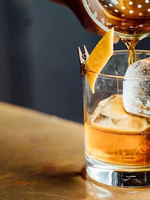 A glass with an orange garnish holds ice, while a beverage is strained into it from above, creating a classic cocktail presentation.