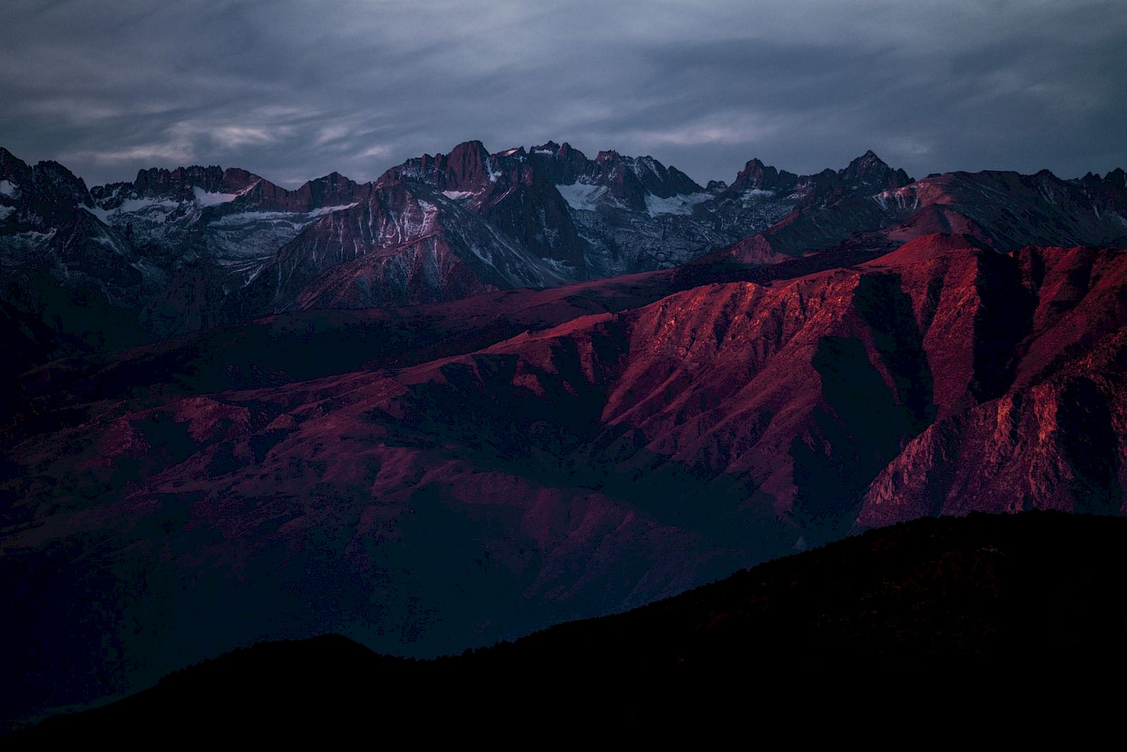 A mountainous landscape at sunset with dramatic red and purple hues illuminating the rugged terrain below a cloudy sky.