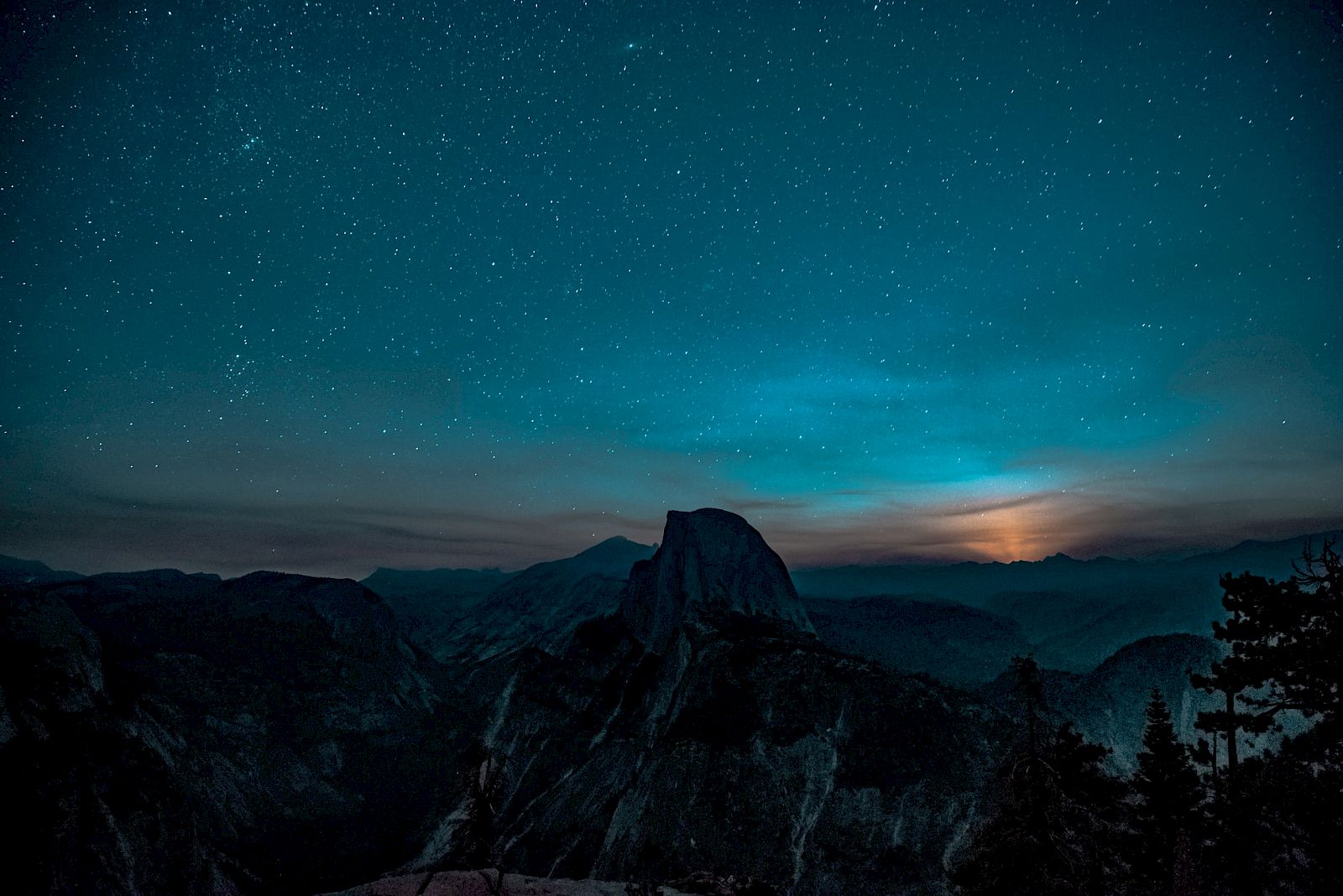 A starry night sky over a mountainous landscape with a glowing horizon and silhouetted trees in the foreground.