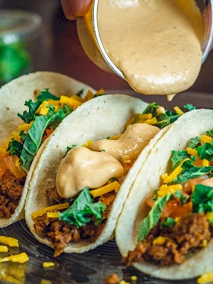 Three tacos filled with meat, cheese, lettuce, and tomatoes, with a sauce being poured on top.