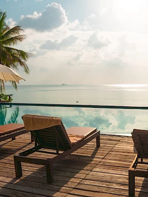 Poolside deck with lounge chairs and umbrellas overlooking a calm sea, surrounded by palm trees under a partly cloudy sky.