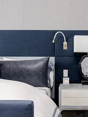 A modern bedroom features a bed with blue and white bedding, a wall-mounted reading light, and a nightstand with a lamp and decorations.