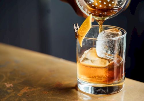 A cocktail is being poured into a glass with ice and a lemon twist garnish on a table.