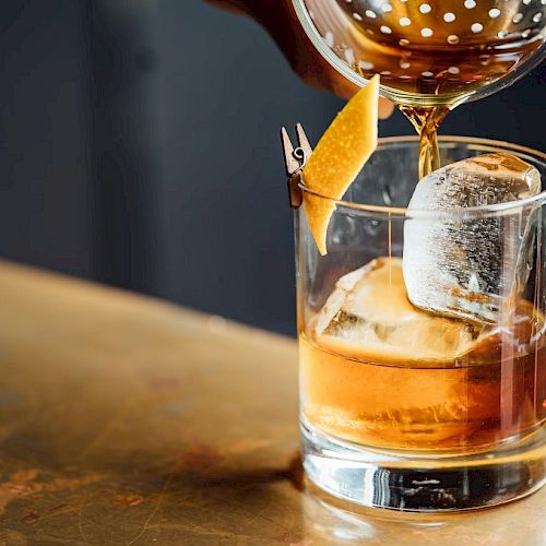 A cocktail is being poured into a glass with ice and a lemon twist garnish on a table.