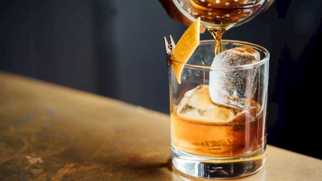 A cocktail is being poured into a glass with ice and a lemon twist garnish on a table.