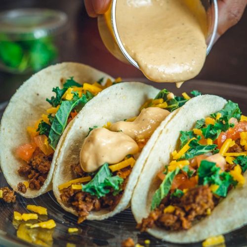 Three tacos filled with ground meat, cheese, lettuce, and tomatoes, topped with sauce being poured over them.