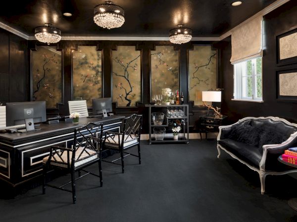 A stylish office with a black desk, three chairs, computers, floral artwork, elegant sofas, and a drink cart.