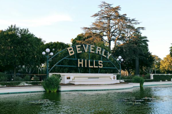 The image shows the iconic "Beverly Hills" sign above a green pond, surrounded by lush trees and walkway lights.