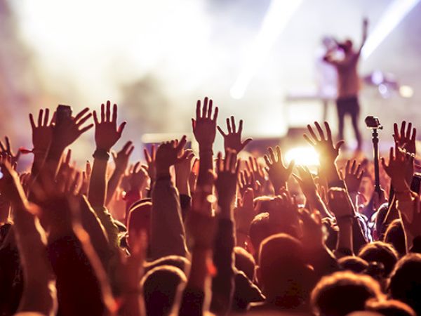 A crowd at a concert with raised hands, facing a brightly lit stage where a performer is singing under spotlights.