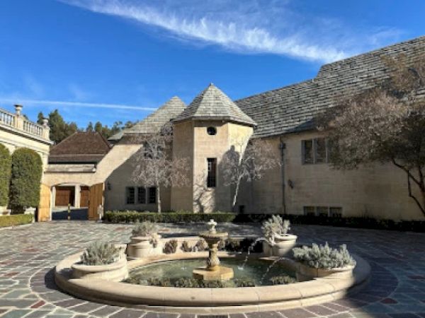 A stone courtyard features a central fountain, surrounded by trees and a large building with gray shingles under a clear blue sky.