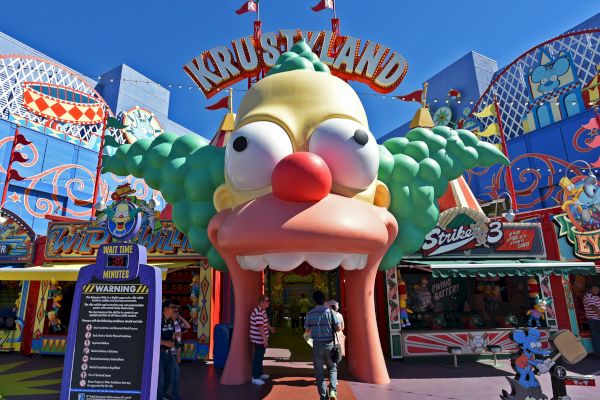 An entrance to an amusement park themed as "Krustyland" with a large clown face and colorful signage, including a wait time sign.