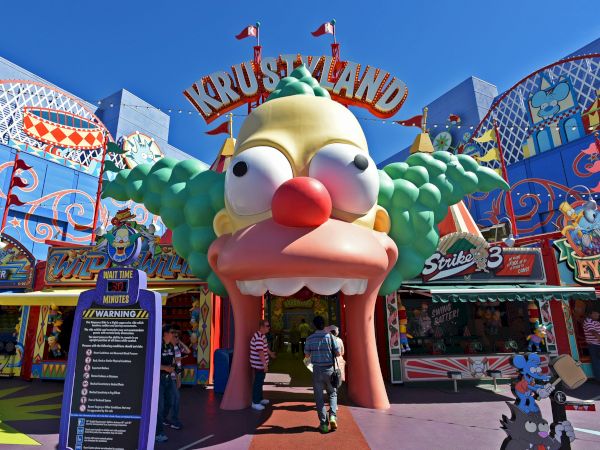 An entrance to an amusement park themed as "Krustyland" with a large clown face and colorful signage, including a wait time sign.