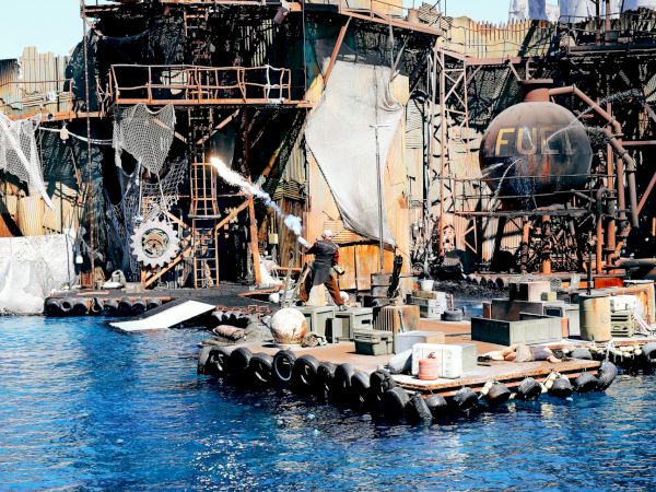 The image shows a water-based action scene with a performer, industrial structures, and a fuel tank, likely from a themed entertainment show.