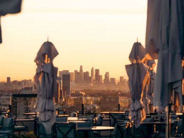 Rooftop terrace with tables and closed umbrellas at sunset, overlooking a city skyline in the background.