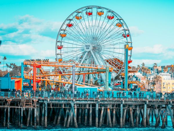 The image shows a lively pier with a Ferris wheel, other amusement rides, and people enjoying the seaside atmosphere under a blue sky.