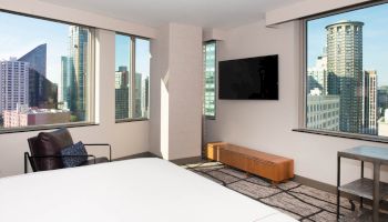 A modern hotel room with a large bed, city views through windows, a TV, and minimalistic furniture.