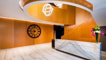 A modern lobby with a marble reception desk, wood accents, a wall clock, and decorative lighting, featuring a flower arrangement.