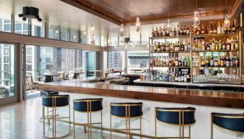 A modern bar with sleek stools, a well-stocked shelf of bottles, large windows, and a stylish ceiling in a high-rise setting.
