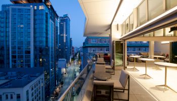 A modern balcony with seating overlooks a city street at dusk, with tall buildings on the left and well-lit indoor space on the right.