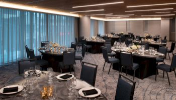 The image shows an elegantly set dining area with round tables, black chairs, place settings, and floral centerpieces, along with dim lighting.