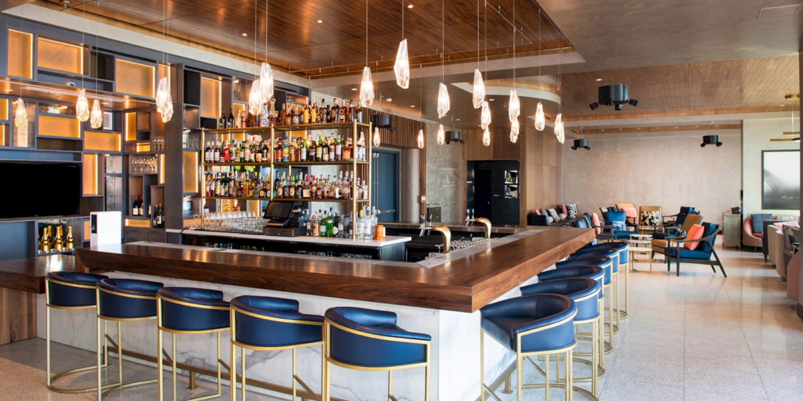 A modern bar with a well-stocked liquor display, surrounded by blue chairs and elegant lighting in a spacious, stylish setting.