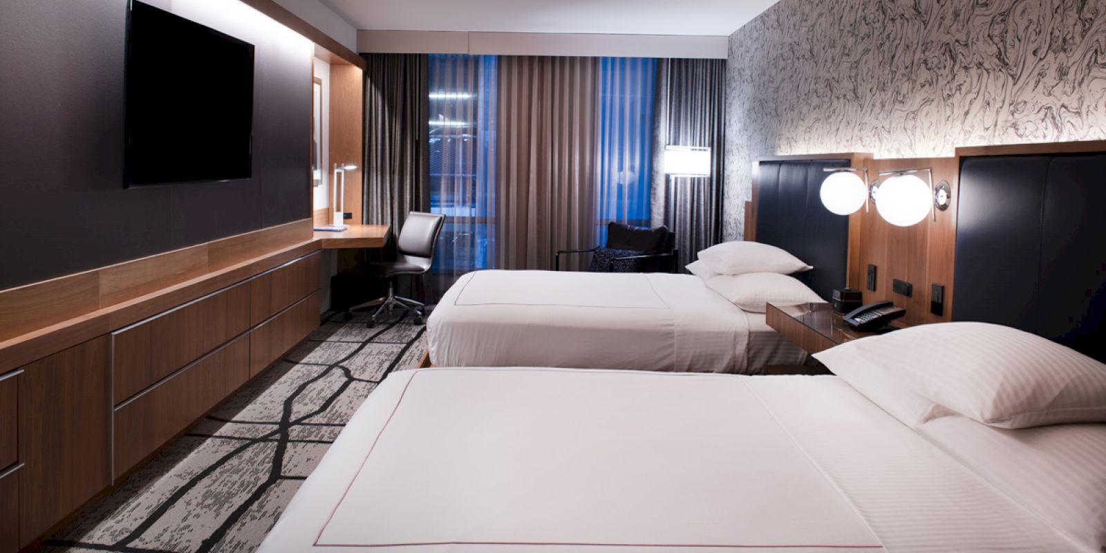 A modern hotel room with two beds, a wall-mounted TV, a desk with chair, and patterned wallpaper, creating a sleek and cozy atmosphere.