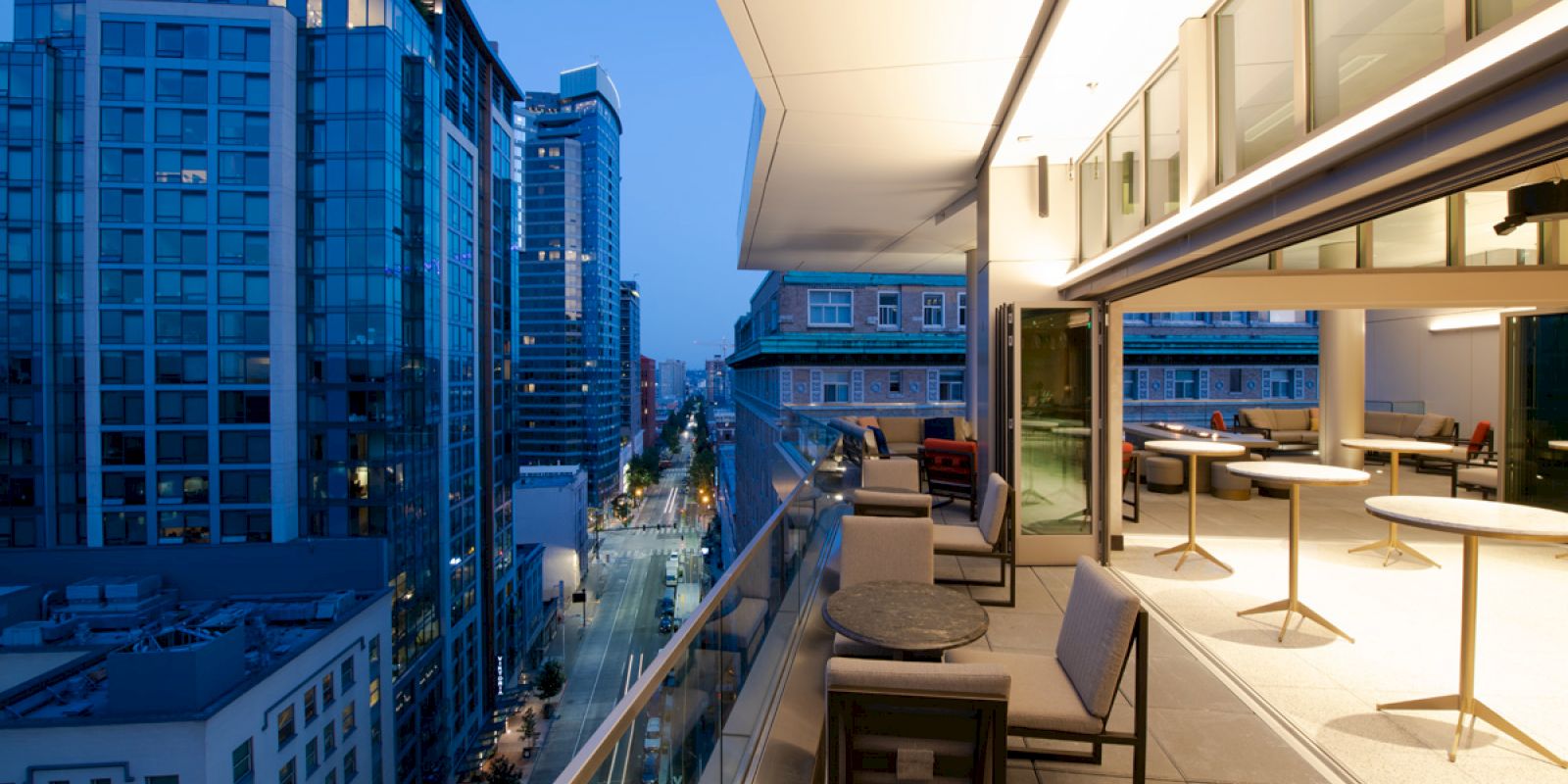 A modern rooftop terrace at dusk with seating and city views, featuring sleek furniture and illuminated buildings in the background.