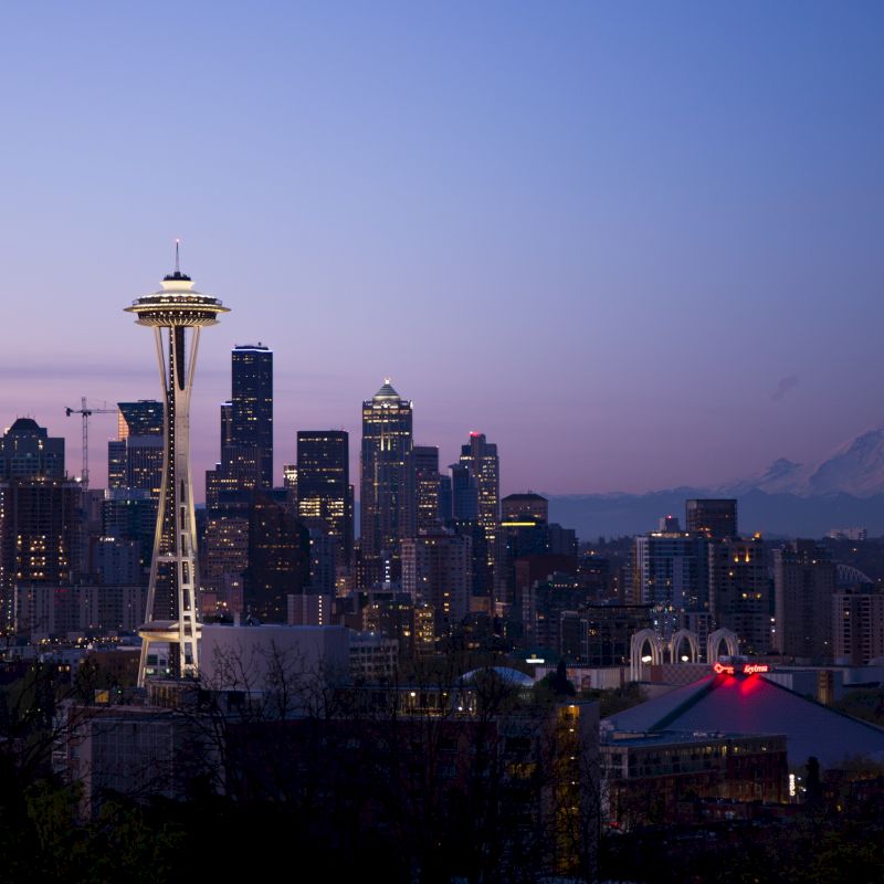 Seattle skyline at twilight, featuring the Space Needle and Mount Rainier in the background. The city lights contrast the evening sky.