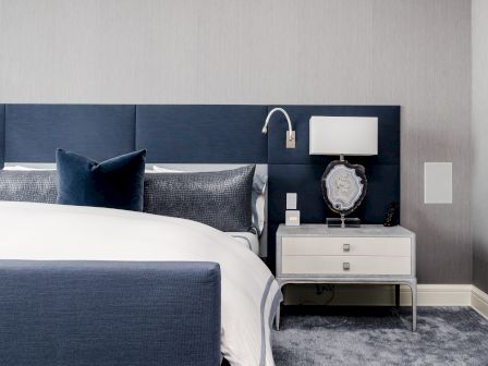 A modern bedroom features a blue upholstered bed, white bedding, a nightstand with a lamp, and a decorative piece on a gray carpeted floor.