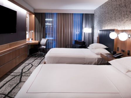 The image shows a modern hotel room with two beds, a desk with a chair, a TV, and decorative lighting on the walls.