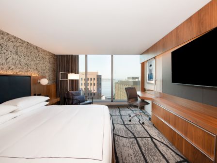 A modern hotel room features a large bed, wall-mounted TV, desk, and a window with a city view.